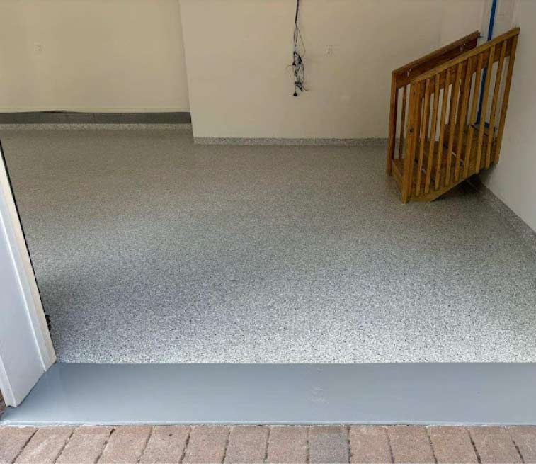Close-up of a professional garage floor coating installation showing the seamless transition from a decorative flake epoxy surface to a solid grey protective threshold near a wooden staircase