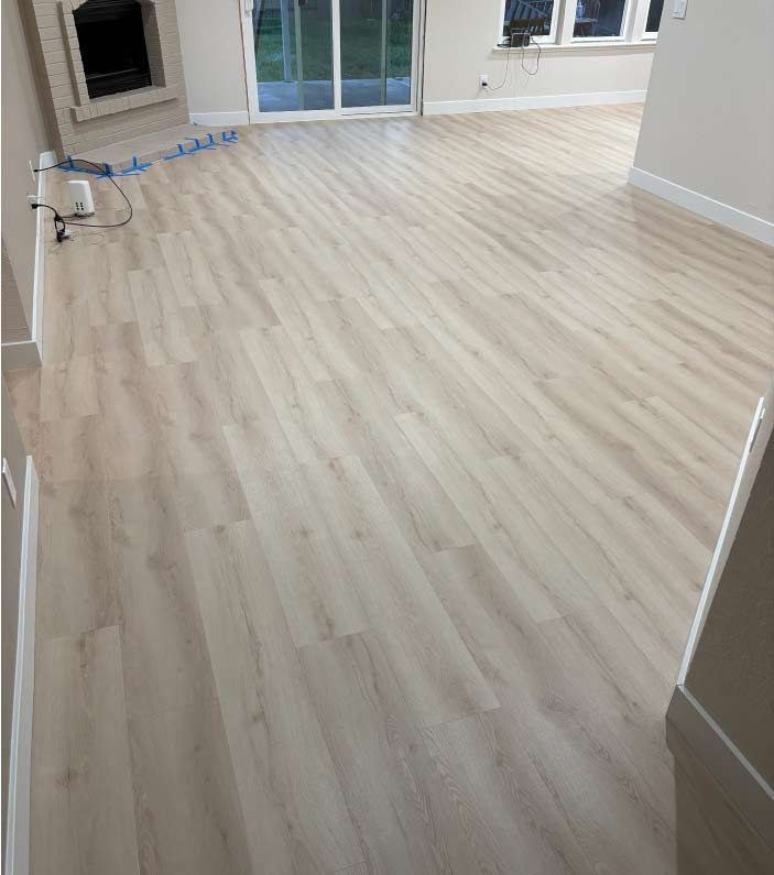 Spacious living room area featuring newly installed luxury vinyl plank (LVP) flooring in a natural wood tone, extending toward a brick fireplace and sliding glass patio doors