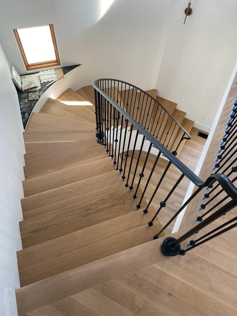 Architectural curved hardwood staircase viewed from above, highlighting premium light oak steps paired with sleek, modern black iron balusters.